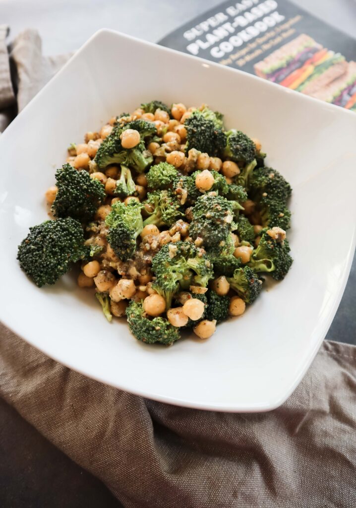 broccoli, walnuts, and chickpeas mixed i white bowl with mustard dressing photographed on cloth next to super easy plant-based cookbook