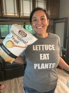 Kathy (woman) wearing a shirt that reads "Lettuce Eat Plants" and holding large bag of Foods Alive Brand Nutritional Yeast 