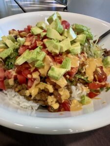 white bowl of white rice, with lentil bulgar taco filling, lettuce, tomatoes, olives, drizzled with cashew queso and chopped avocado