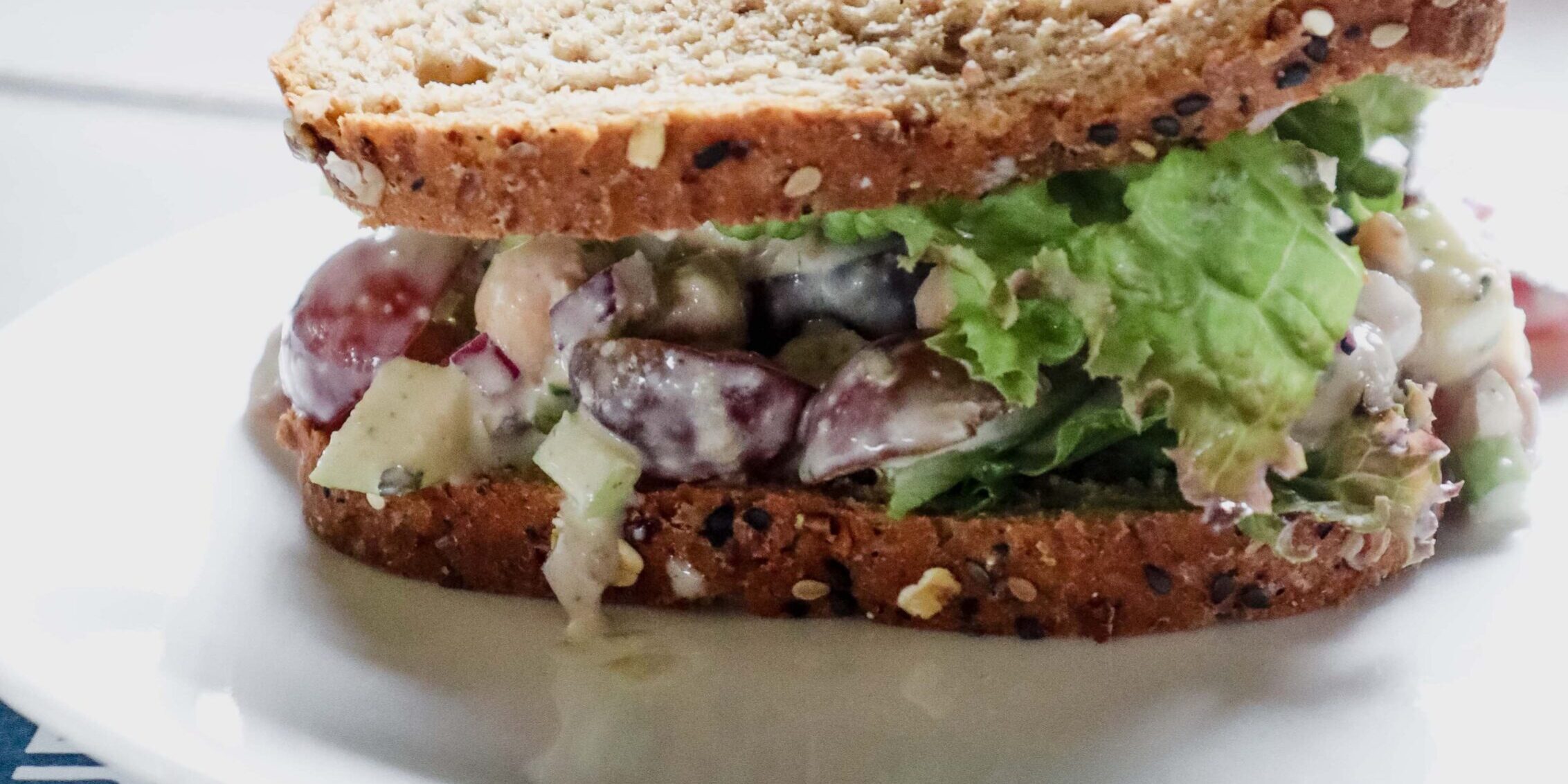 sandwich on whole grain seeded bread with side angle shot, full of a creamy chickpea, grape, apply, and onion salad filling.