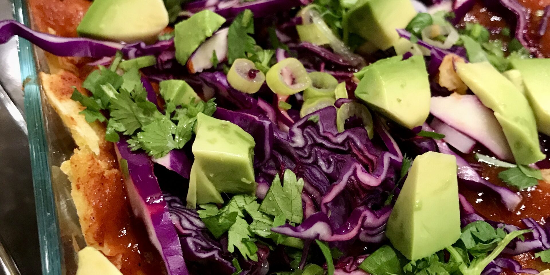 glass baking dish with enchiladas and sauce topped with cabbage, avocado, cilantro, and green onions.