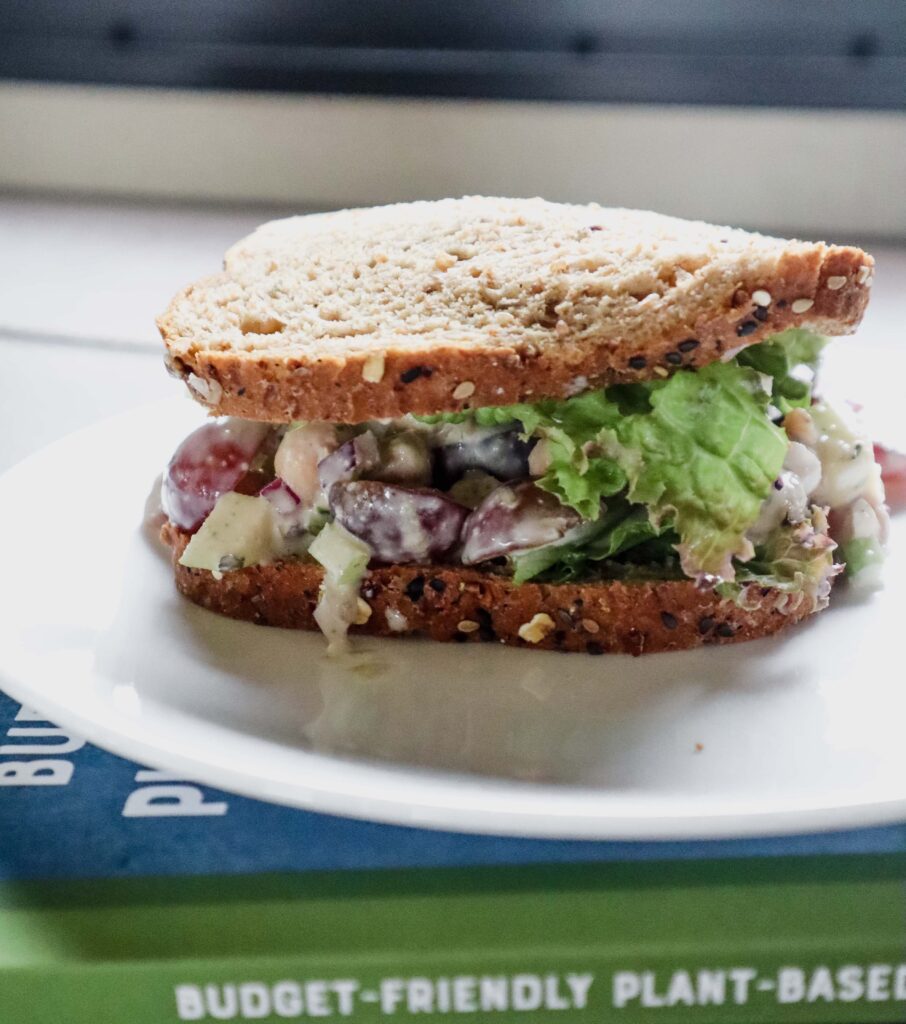 sandwich on whole grain seeded bread with side angle shot, full of a creamy chickpea, grape, apply, and onion salad filling.