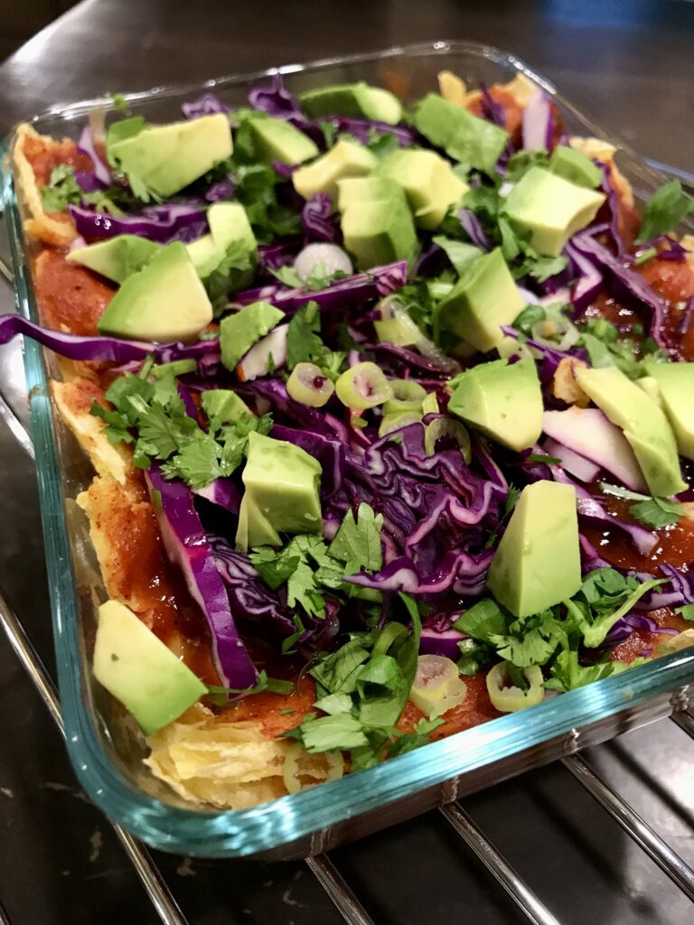 glass baking dish with enchiladas and sauce topped with cabbage, avocado, cilantro, and green onions.