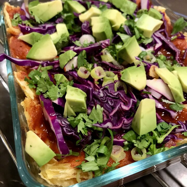 glass baking dish with enchiladas and sauce topped with cabbage, avocado, cilantro, and green onions.